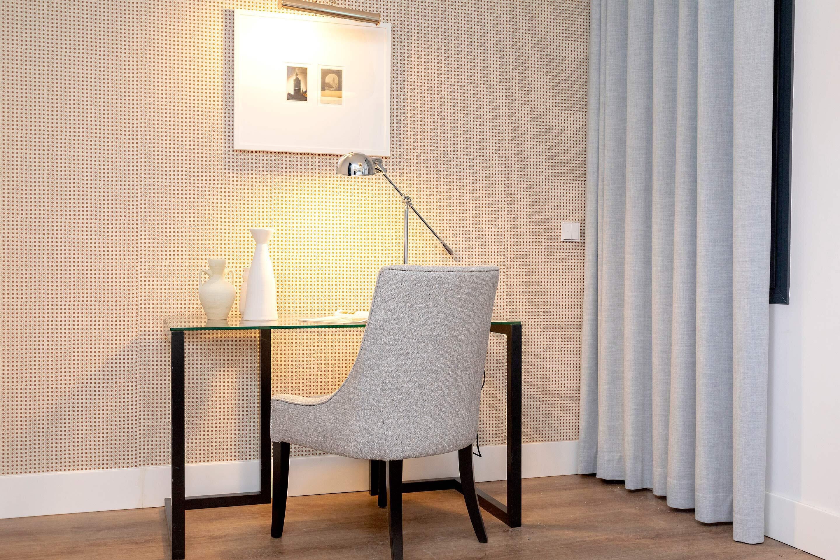 Una habitación moderna con un escritorio de vidrio, una silla gris, un jarrón blanco y un marco de fotos en la pared.