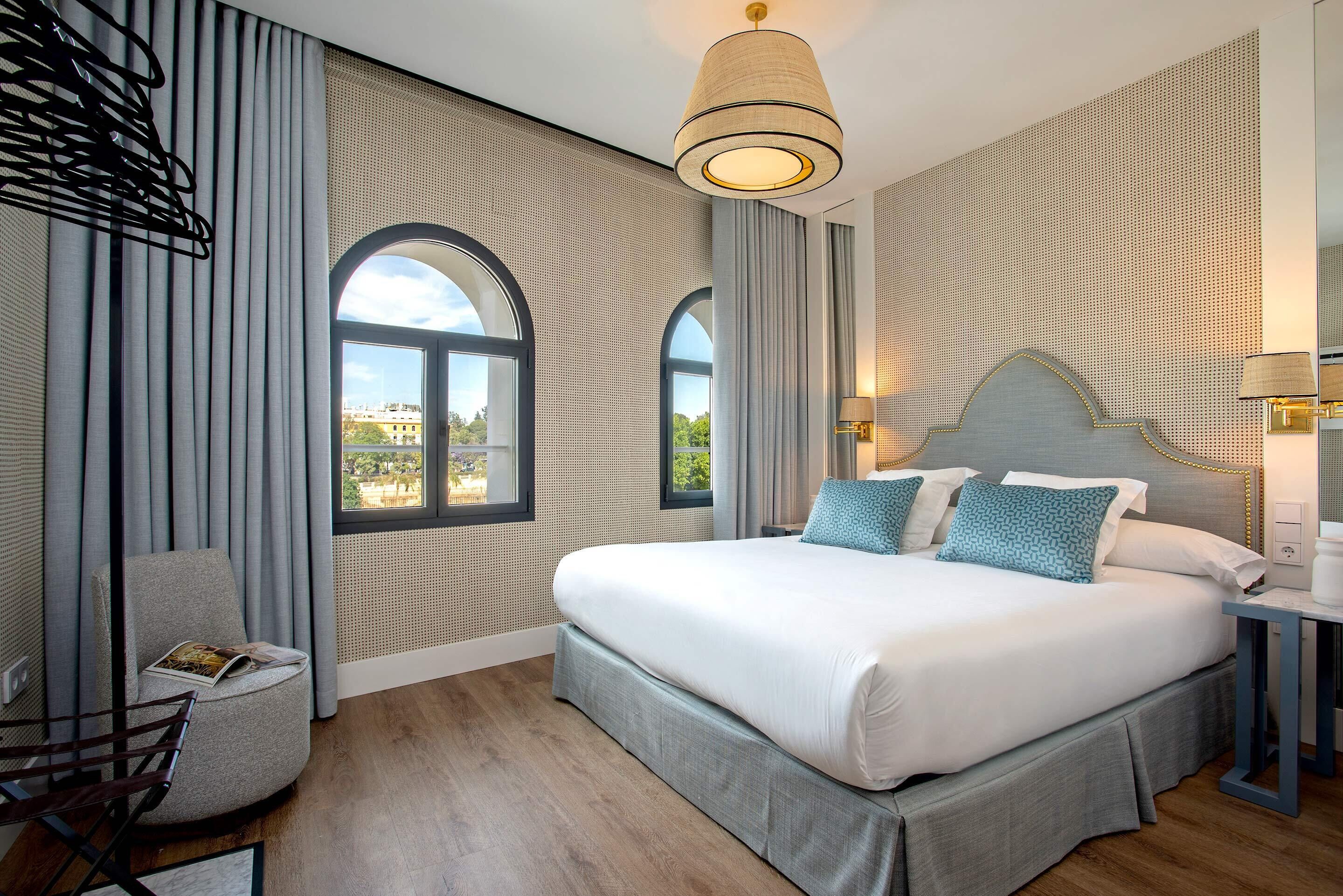 A modern hotel room with a large bed, two windows, a chair, and a lamp.