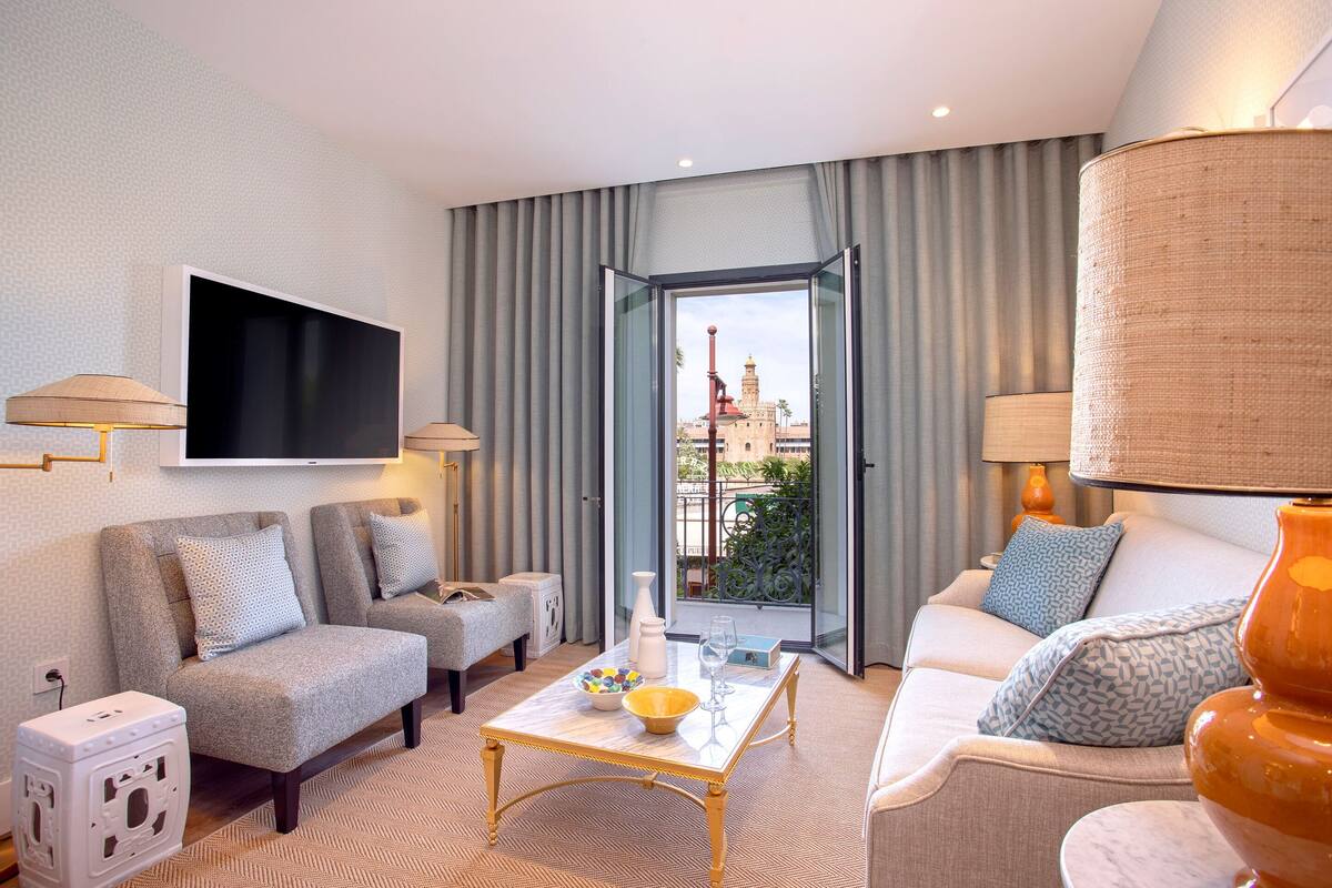 A modern hotel room with a sofa, armchairs, a coffee table, and a TV.