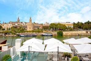 A riverside terrace with outdoor seating and a view of historic buildings and boats. - Genteel Home Betis 67 (Seville)