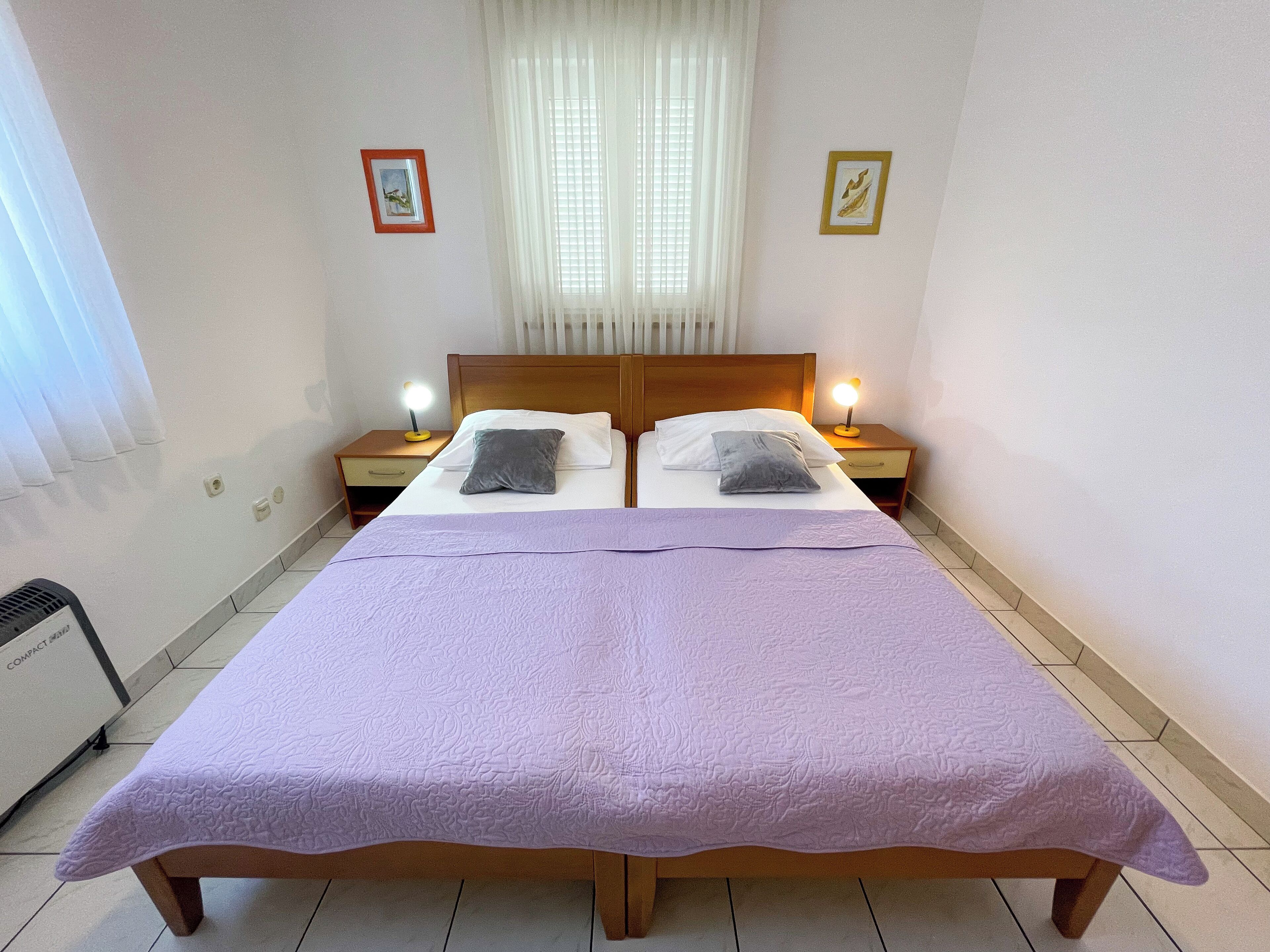 A double bed with a purple bedspread, two bedside lamps, and a window with white curtains.