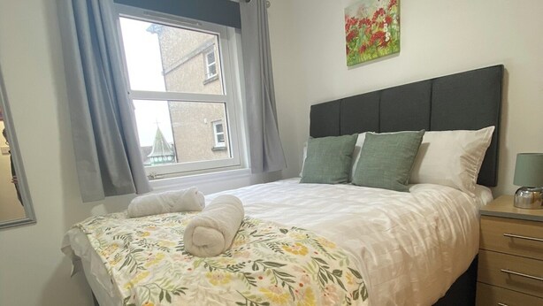 Un lit bien fait, recouvert d’une couvre-lit à motifs floraux, une table de chevet avec une lampe et une fenêtre avec des rideaux.