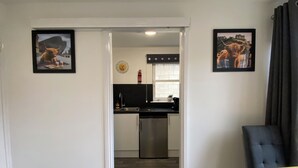 Une cuisine moderne avec un réfrigérateur en acier inoxydable, un plan de travail noir et une fenêtre donnant sur la ville. Des tableaux encadrés ornent les murs, et une chaise bleue se trouve dans l’espace salon.