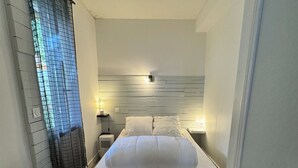 A bedroom with a bed, bedside tables, a nightstand, a wall-mounted light, and a window with a view. - Apart 50m² 5 People 2 Bedrooms –Town, Train, Beach few mn (La Bernerie-en-Retz)