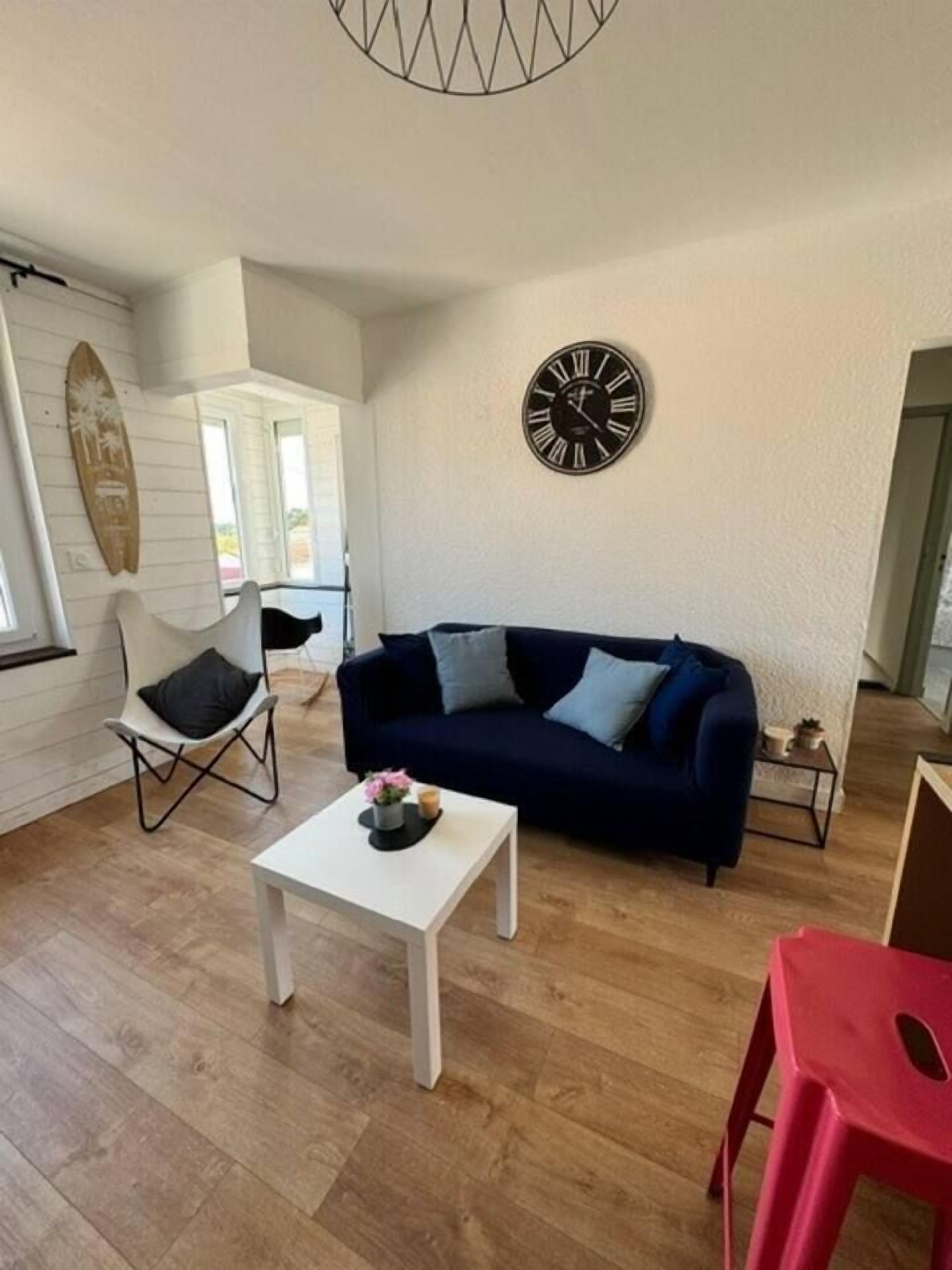 Un salon avec un canapé bleu foncé, une table basse blanche, un fauteuil rouge et une horloge murale.
