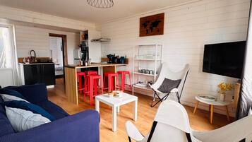 Un salon moderne avec un canapé, une table basse et des fauteuils, un coin cuisine avec des tabourets rouges et un téléviseur fixé au mur.
