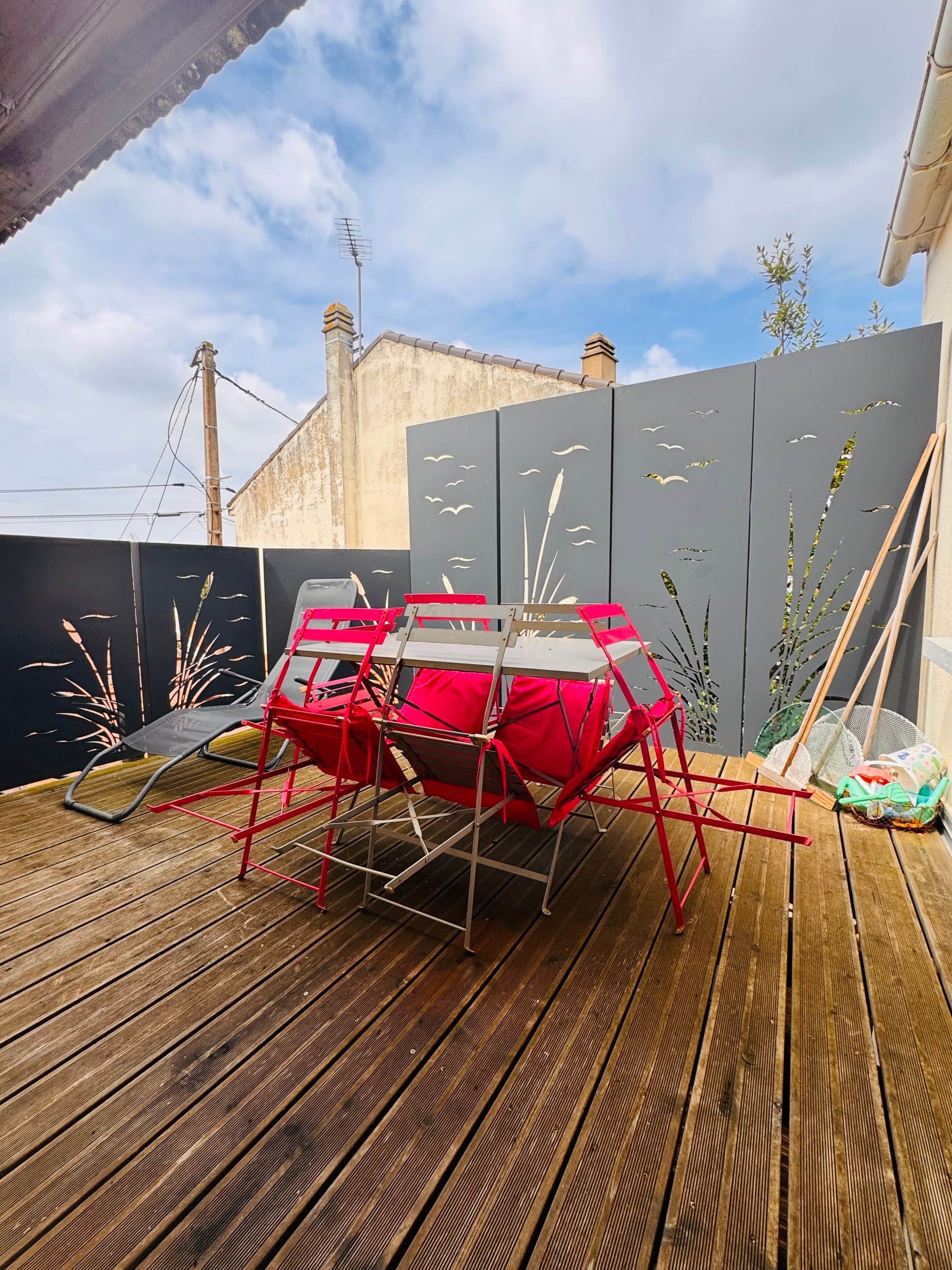 Une terrasse en bois avec une chaise rouge et une table, une clôture grise ornée de motifs découpés, et un bâtiment en arrière-plan.