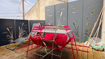 Une terrasse en bois avec une chaise rouge et une table, une clôture grise ornée de motifs découpés, et un bâtiment en arrière-plan.