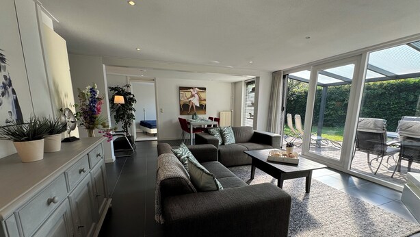 A modern living room with a sofa, coffee table, and dining area. There is a large sliding glass door leading to an outdoor patio. - Detached 4-person house with sauna, WLAN, fenced garden. (Brouwershaven)