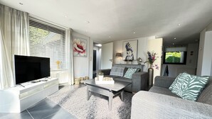 A modern living room with a flat-screen TV, grey sofas, a coffee table, and artwork on the walls. - Detached 4-person house with sauna, WLAN, fenced garden. (Brouwershaven)