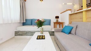 A modern living room with a sofa, a coffee table with a vase and cookies, a bed with blue pillows, and a small wooden table with a lamp.