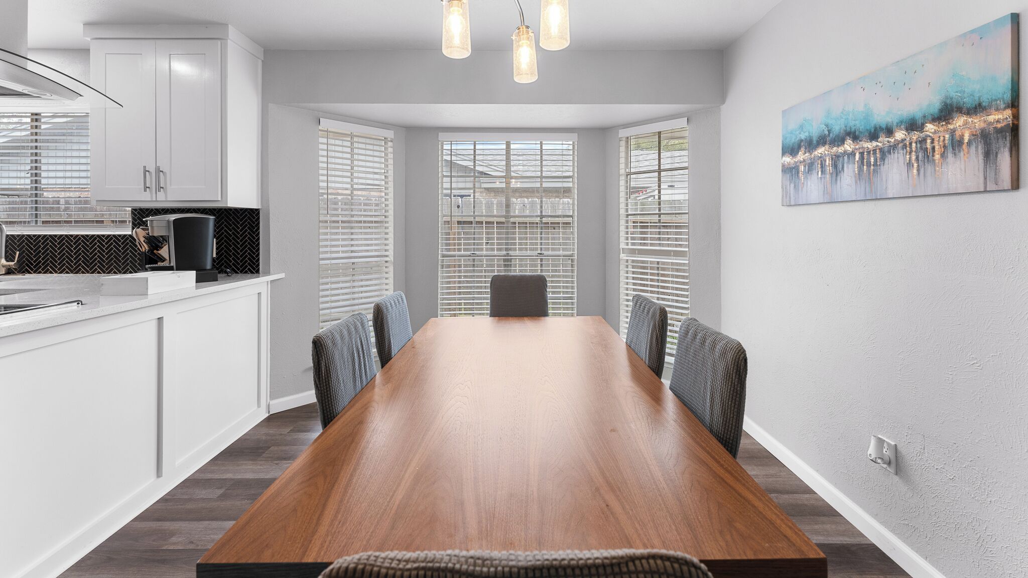 Een moderne keuken met witte kasten, een houten eettafel en een groot raam met zonwering.