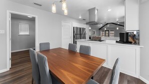 Una cocina moderna con armarios blancos, una isla central, electrodomésticos de acero inoxidable y un comedor con mesa de madera y sillas tapizadas en gris.