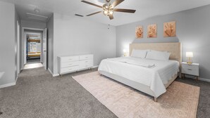 Un dormitorio con cama, mesita de noche, cómoda, ventilador de techo y puerta que da a otra habitación.