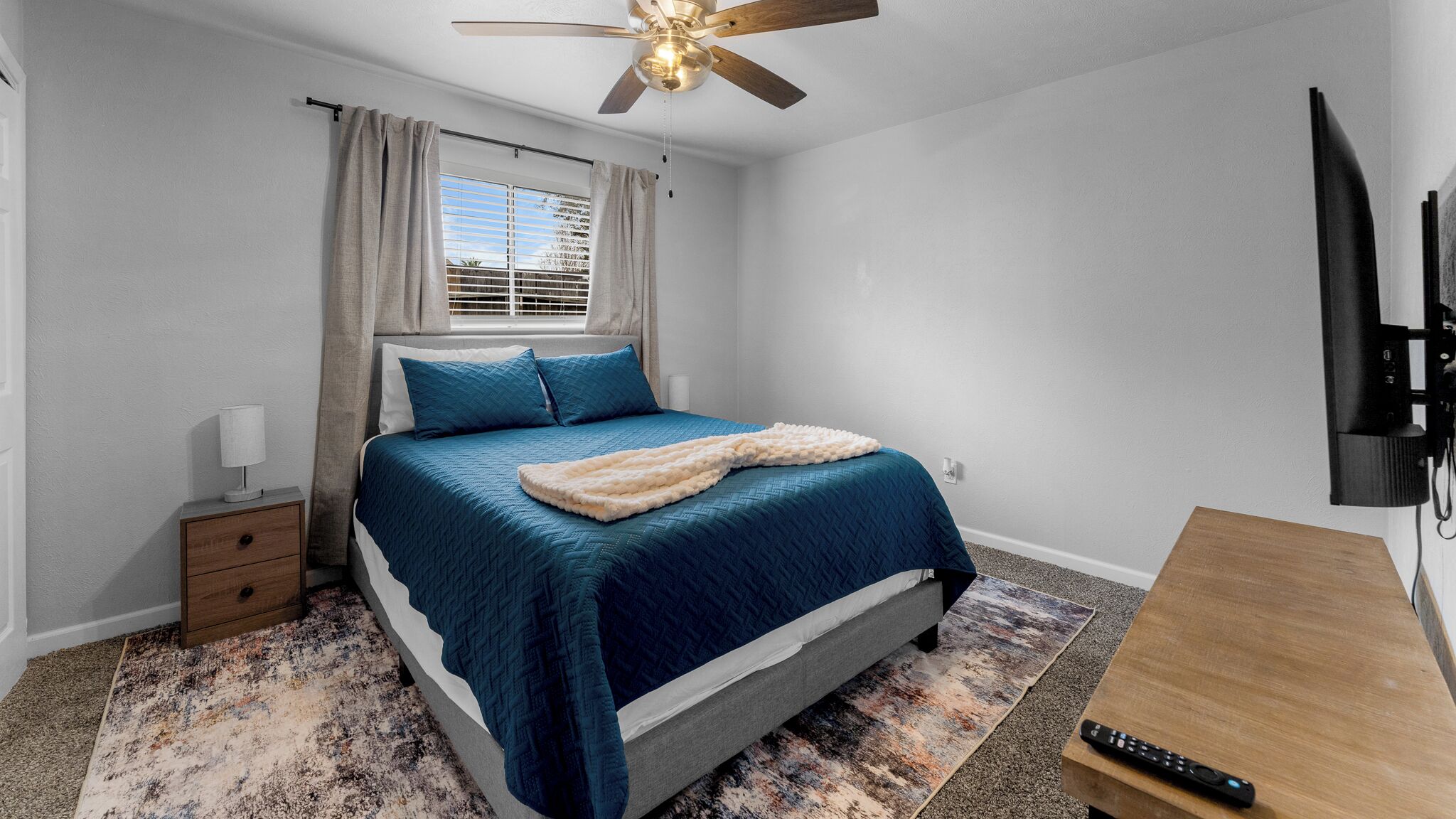 A bedroom with a bed, a nightstand, a desk, a television, and a ceiling fan.