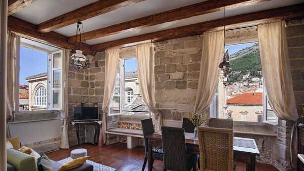 A cozy room with stone walls, wooden beams, a dining area with wicker chairs, and a view of a town with red-roofed buildings.