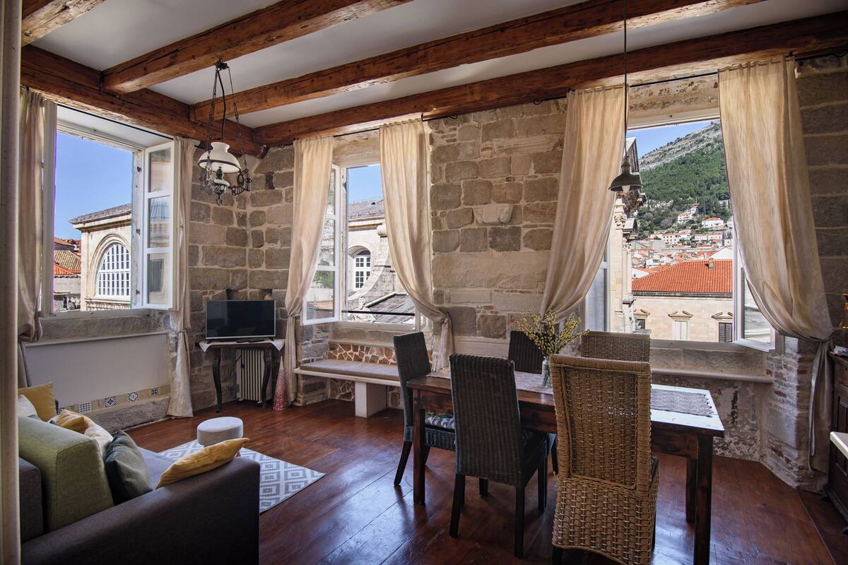 A cozy room with stone walls, wooden beams, a dining area with wicker chairs, and a view of a town with red-roofed buildings.