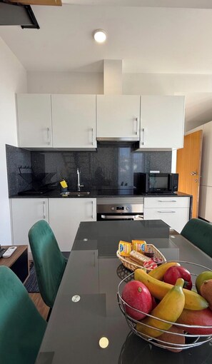 Une cuisine moderne avec des meubles blancs, un plan de travail noir et un coin repas comprenant une table en verre et des chaises vertes.