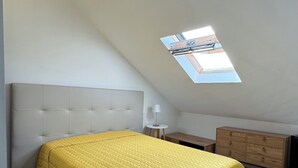 A bedroom with a sloped ceiling, a skylight, a bed with a yellow quilt, a bedside table, and a wooden dresser.