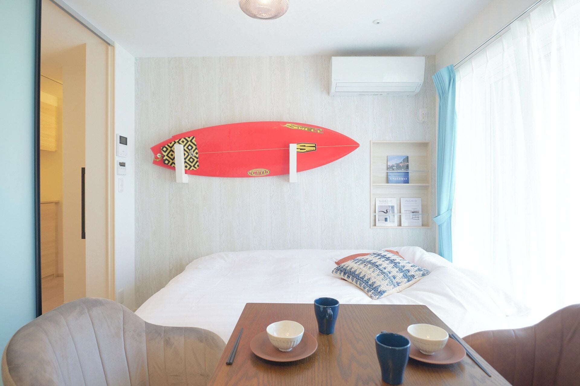 Een hotelkamer met een bed, een tafel met kommen en bekers, een surfplank aan de muur en een plank met boeken.