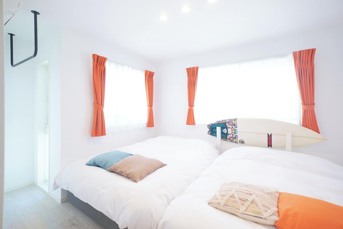 Une chambre avec deux lits, des rideaux orange, une planche de surf et une fenêtre.