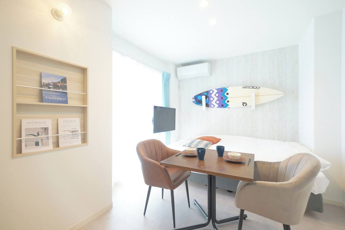 Une chambre d’hôtel comprenant un lit, un bureau avec deux chaises, une planche de surf accrochée au mur, une télévision et une étagère avec des livres.