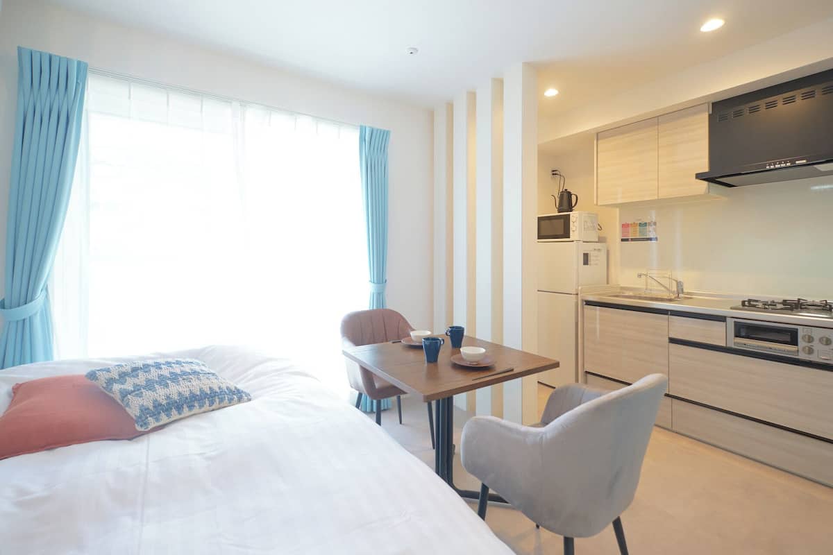 Une chambre d’hôtel comprenant un lit, une table avec des chaises, un coin cuisine et une fenêtre avec des rideaux.