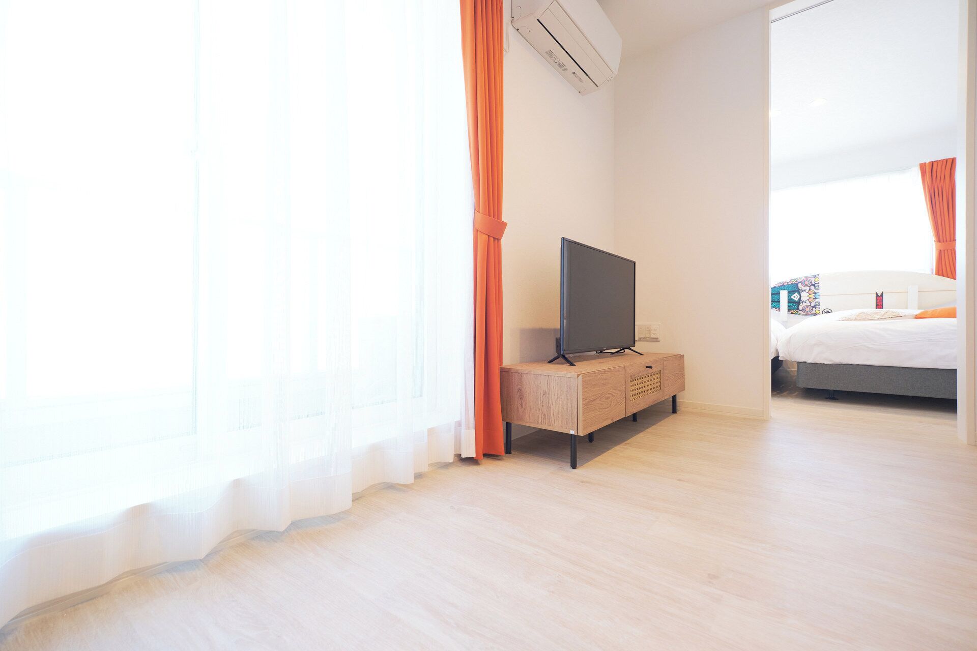 A hotel room with a television on a wooden stand, a bed with a patterned headboard, and a large window with sheer curtains.