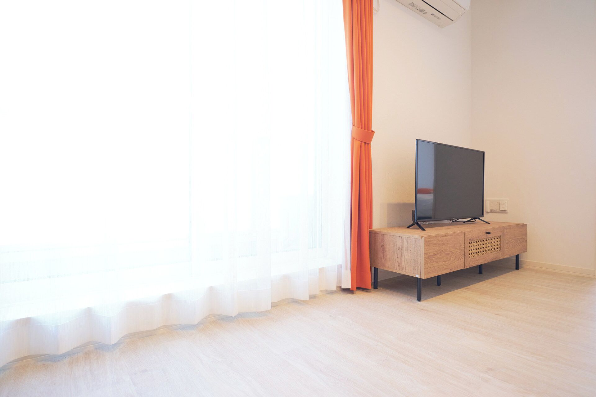 Una habitación con una televisión sobre un mueble de madera, una ventana con cortinas ligeras y un equipo de aire acondicionado en la pared.