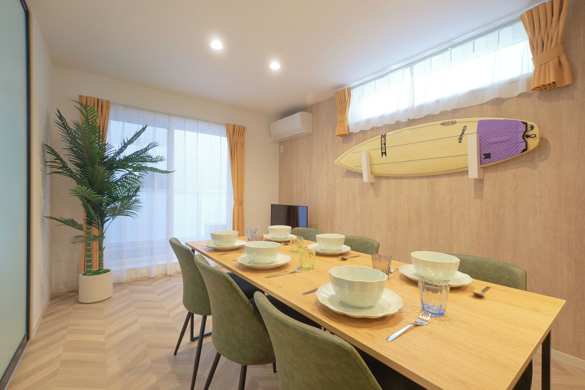 A dining area with a long table set for six, surrounded by chairs. A surfboard is mounted on the wall, and a potted plant is visible in the corner.