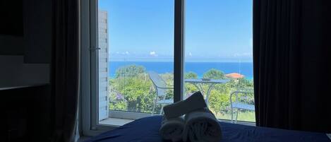Ein Hotelzimmer mit einem Bett, einem Fenster mit Blick auf das Meer und Grünflächen, sowie ein Handtuch auf dem Bett.