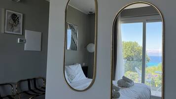 A bedroom with a bed, two mirrors, a bedside table, a chair, and a view of the sea.