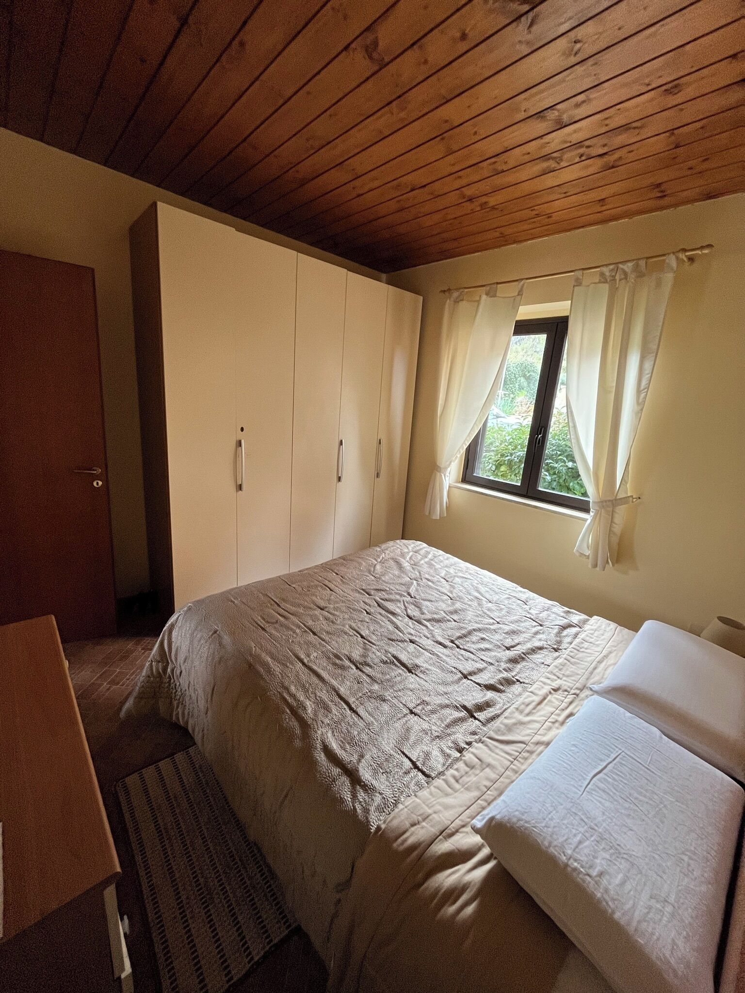 A bedroom with a bed, a wardrobe, a desk, and a window with curtains.