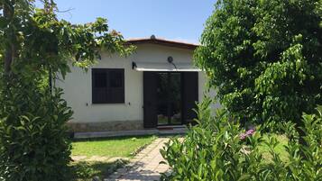 Una casa pequeña de un solo piso con un porche techado, una puerta, una ventana con contraventanas y un acceso con tejas.