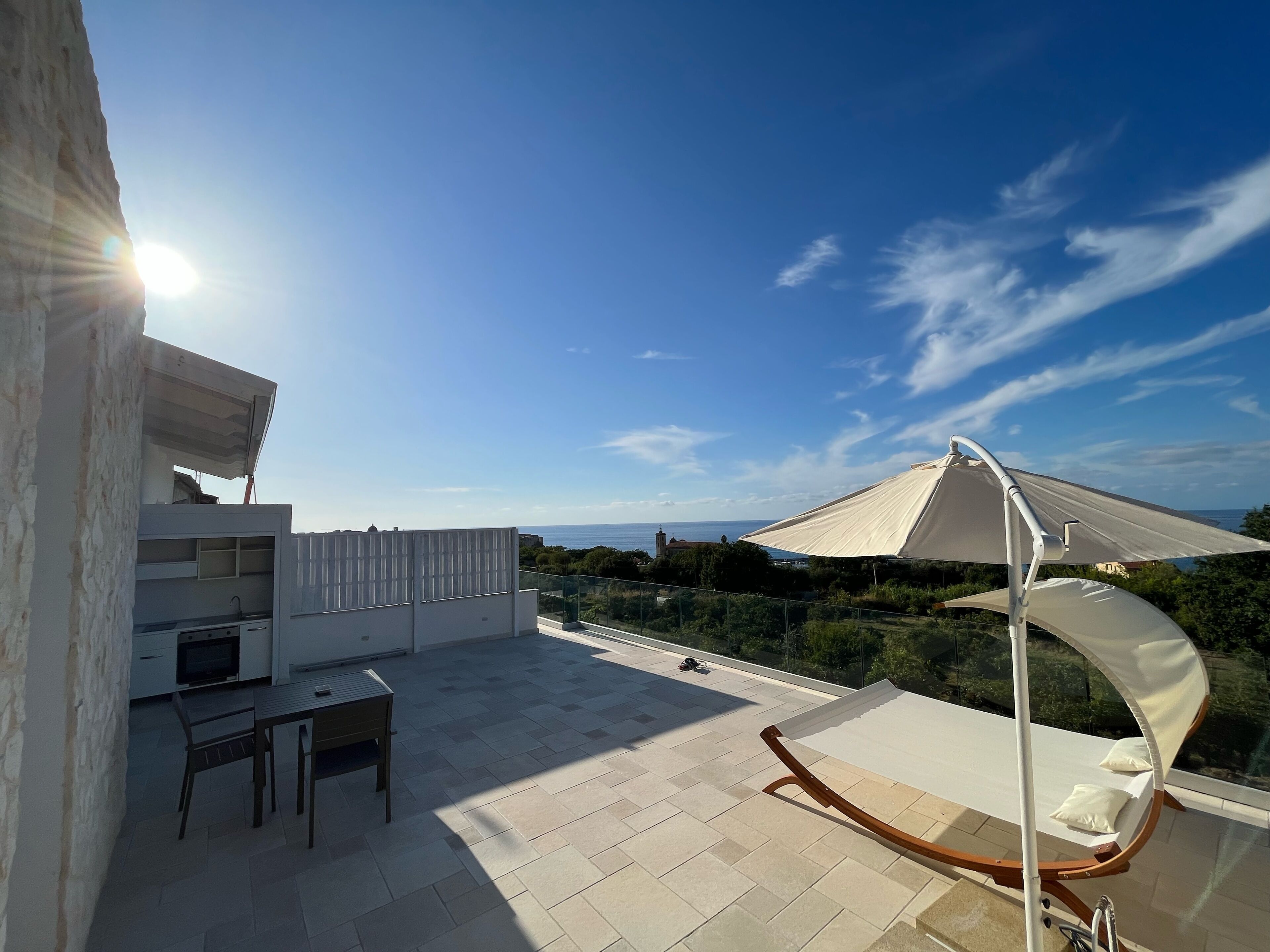 A rooftop terrace with a table and chairs, a white umbrella, and a view of the sea.