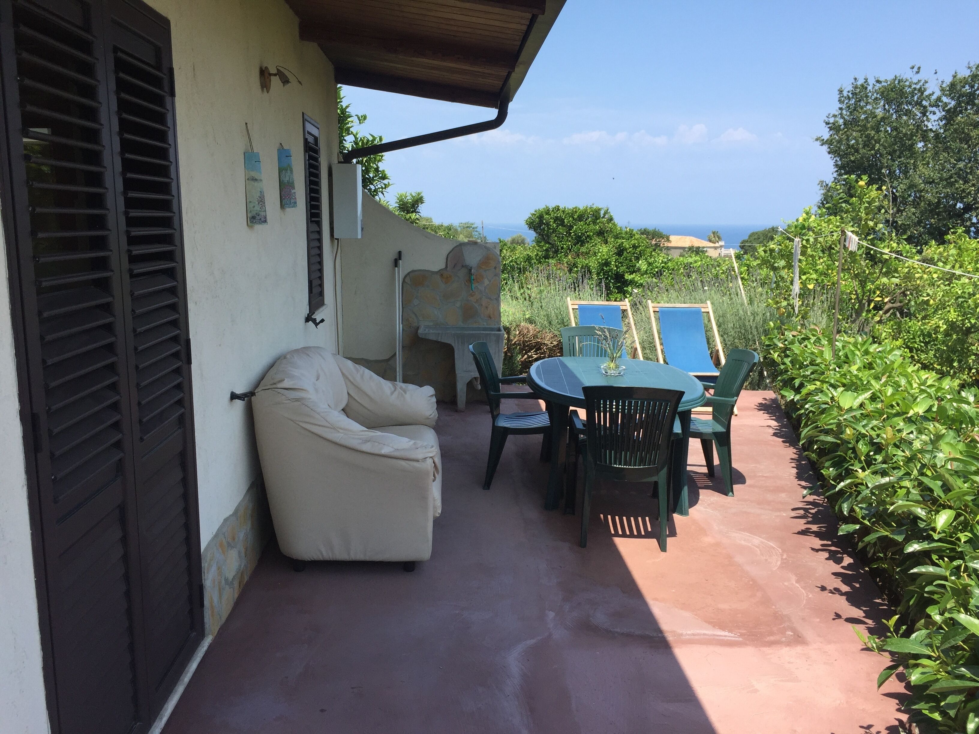 A patio with a beige sofa, a table, and chairs, overlooking a garden and the sea.