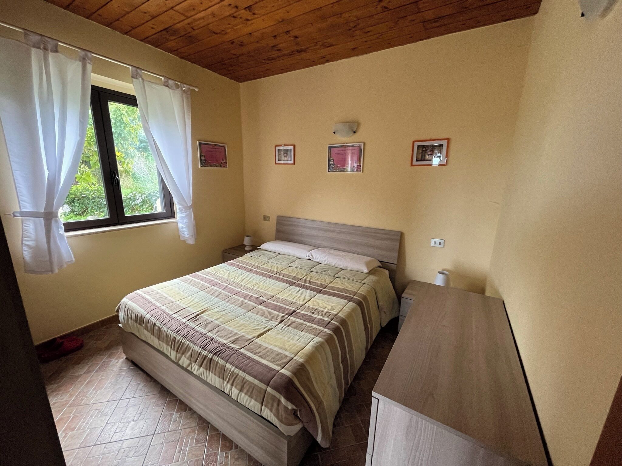 Un dormitorio con cama, mesita de noche, cómoda, ventana con cortinas y cuadros en la pared.