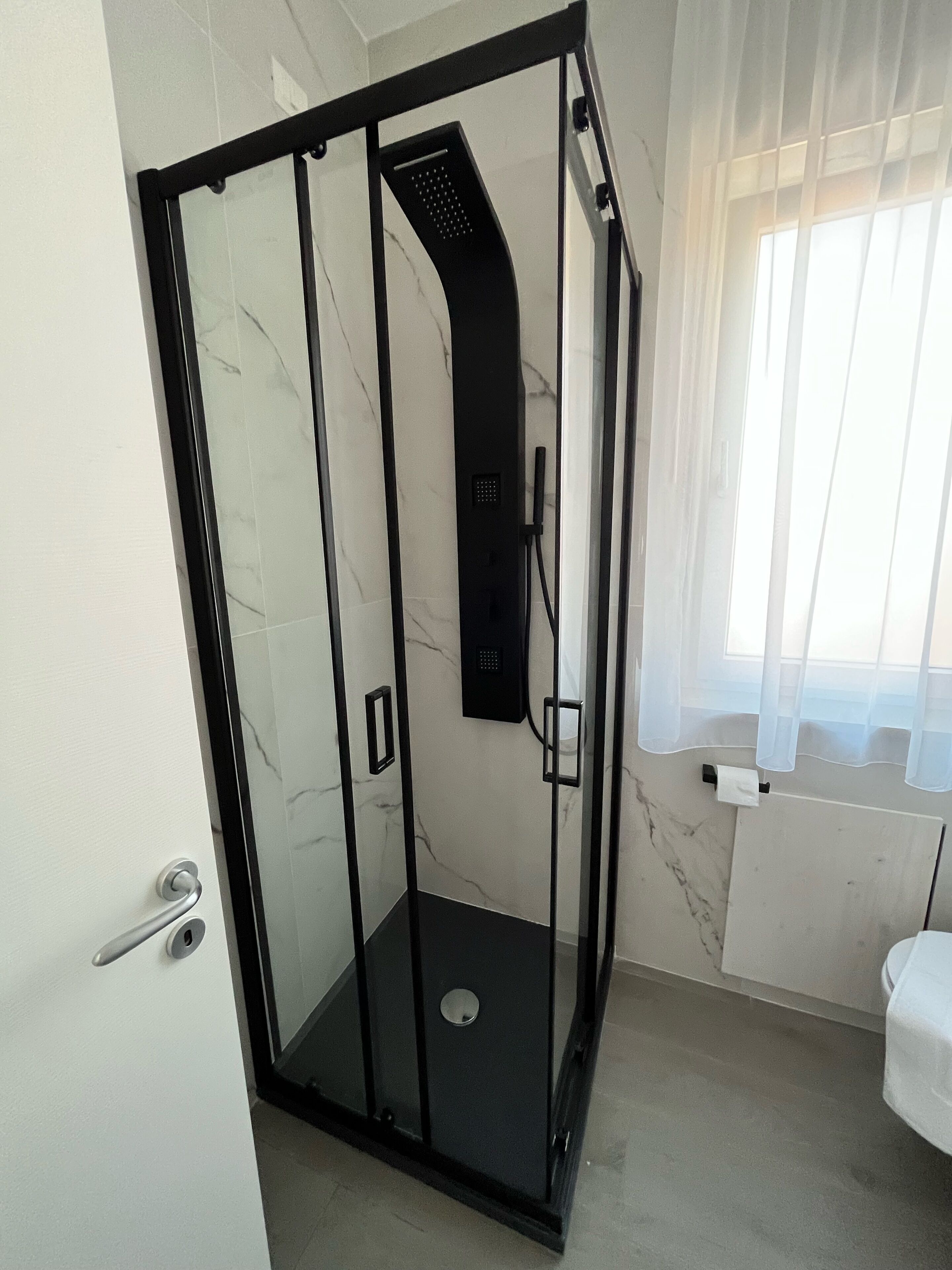 A modern bathroom with a glass-enclosed shower, a toilet, and a window with sheer curtains.