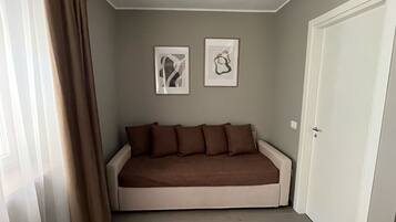 Een kamer met een beige bank, twee kunstwerken in lijsten aan de muur, een witte deur en een raam met gordijnen.