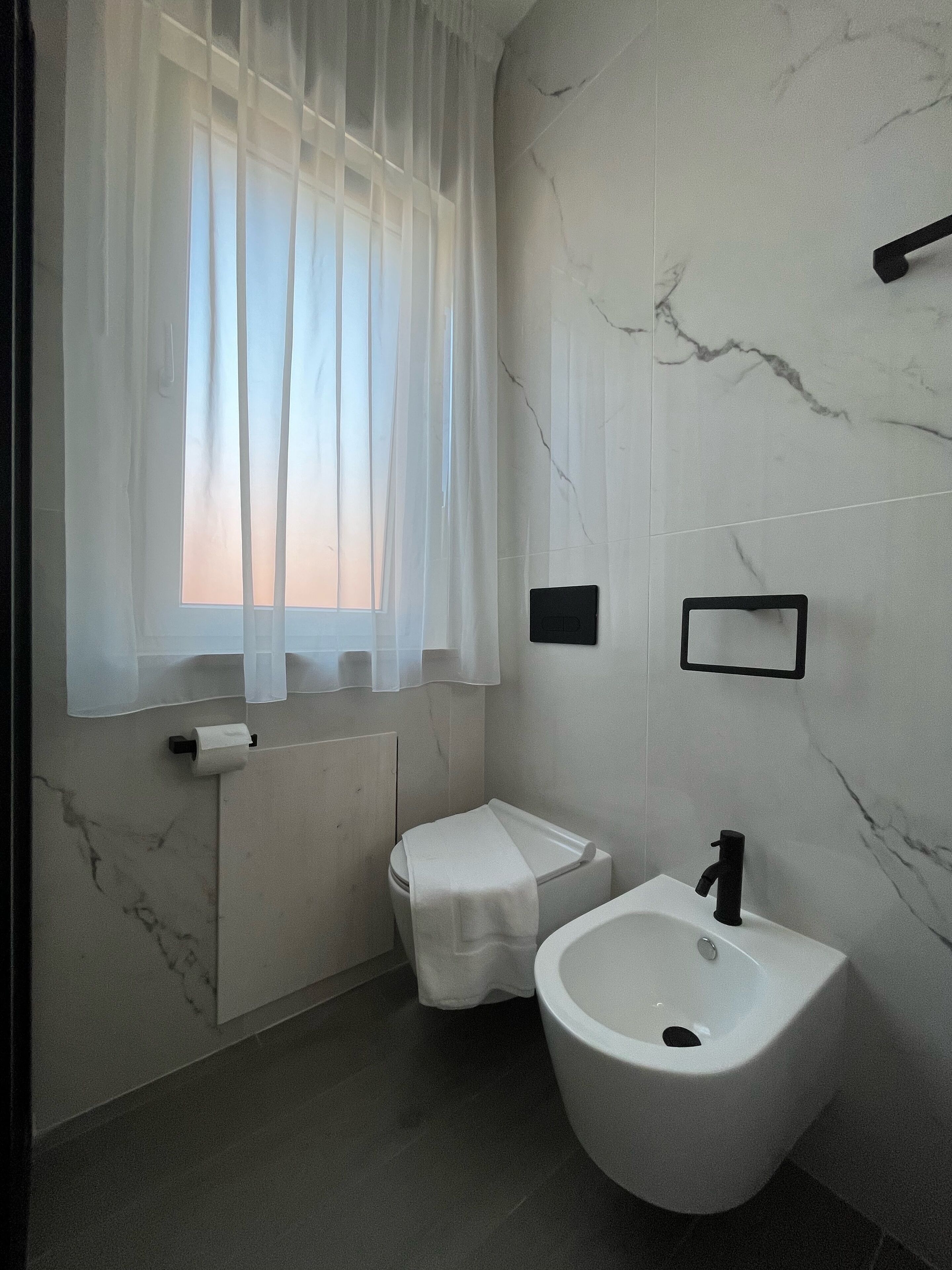 A bathroom with a toilet, bidet, and sink, marble walls, and a window with sheer curtains.