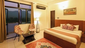 A hotel room with a large bed, two chairs, a small table, a lamp, and a view of mountains through the window. - Hotel Amarah (Jaipur)