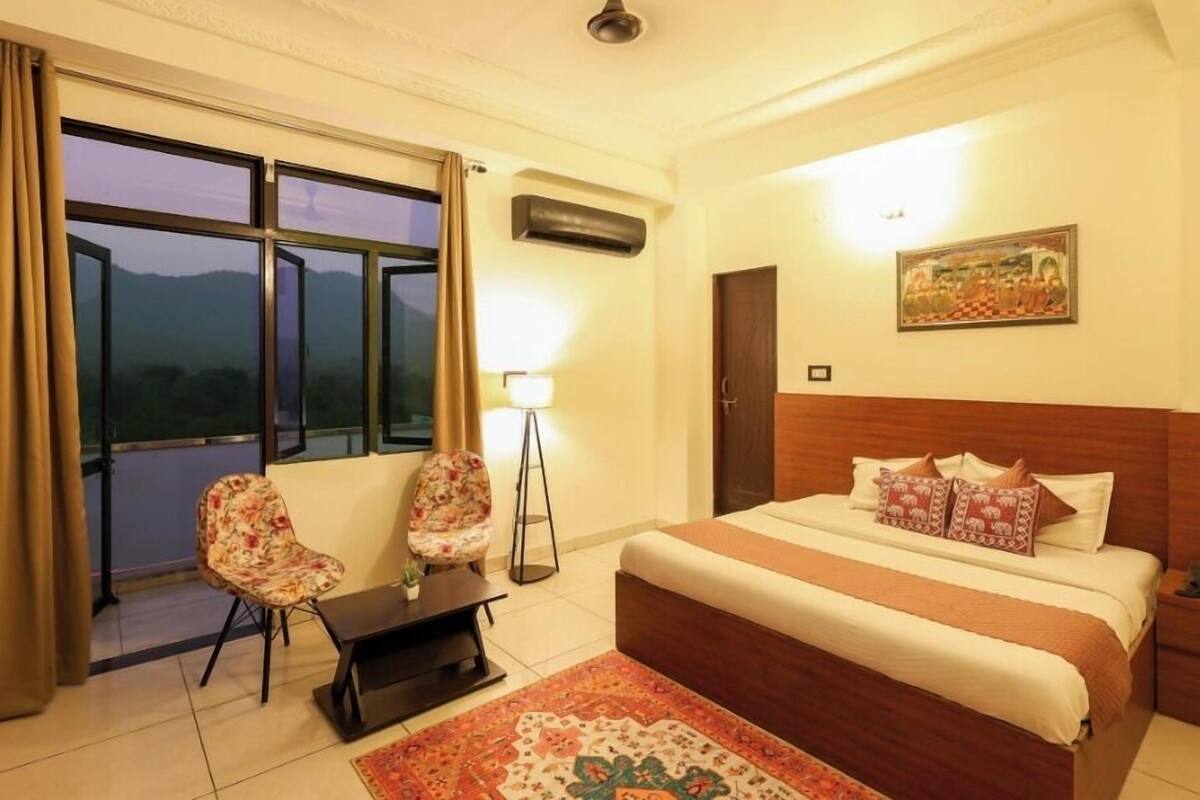 Habitación de hotel con una cama grande, dos sillas, una mesita, una lámpara y vistas a las montañas desde la ventana.
