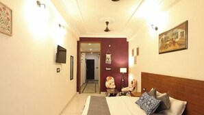 A hotel room with a large bed, a flat-screen TV mounted on the wall, a desk with a chair, and a door leading to another room. - Hotel Amarah (Jaipur)