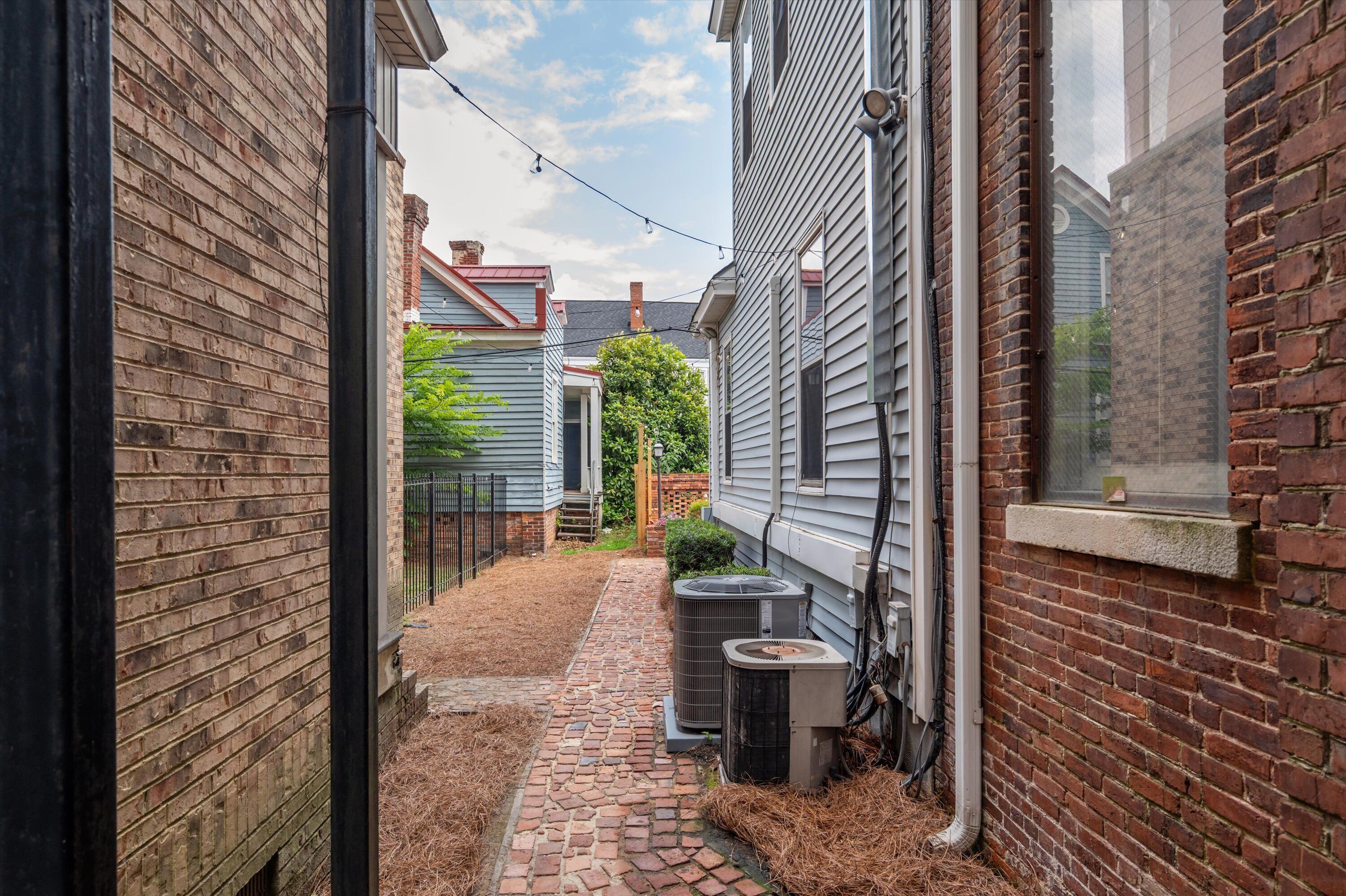 A narrow brick alleyway with a brick pathway, two air conditioning units, and a small window on the right side.