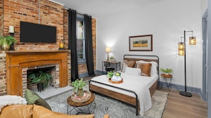 Een moderne slaapkamer met een open haard van baksteen, een bed met houten hoofdeinde, een flatscreen televisie aan de muur en een vloerlamp.