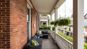 A covered porch with a brick wall, a black sofa with cushions, a small table, and hanging plants. - 1 bedroom, 1 bathroom suite in Downtown Augusta. The sleeping arrangements for this unit are a full bed and a pull-out couch in the living room. (Augusta)