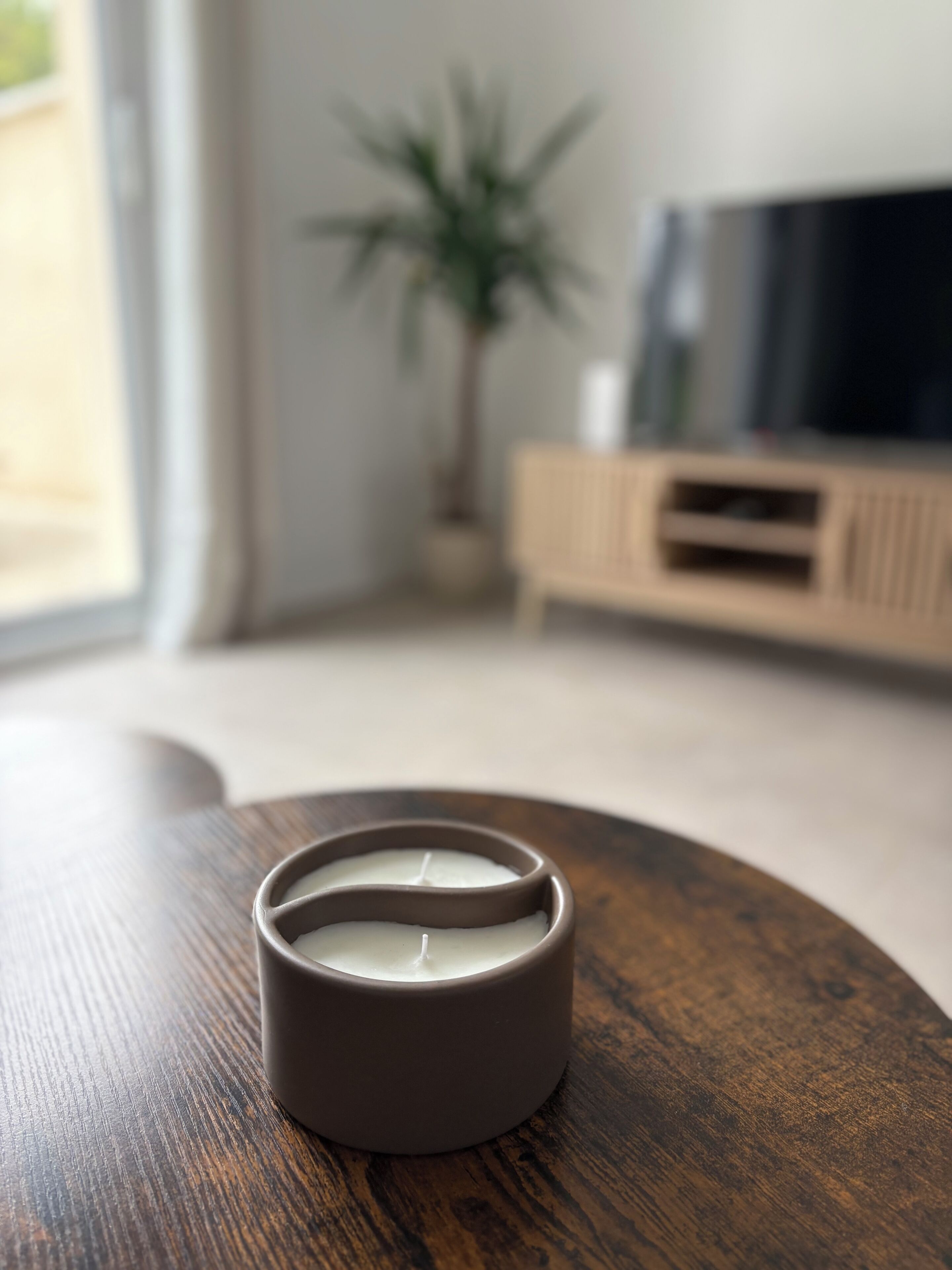 Een kaars in een keramieken houder op een houten tafel, met een vage achtergrond met een televisie en een plantje.