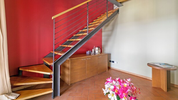 A modern interior with a wooden staircase, a red wall, a wooden cabinet, and a small table with a magazine. - Almavite Wine Resort (Desenzano del Garda)