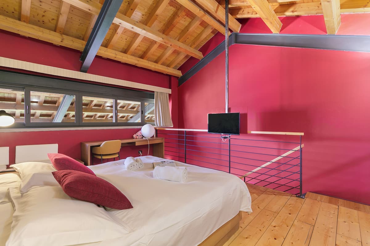 Un dormitorio con techo de madera, pared roja, cama con ropa de cama blanca, escritorio con silla y televisor montado en la pared.