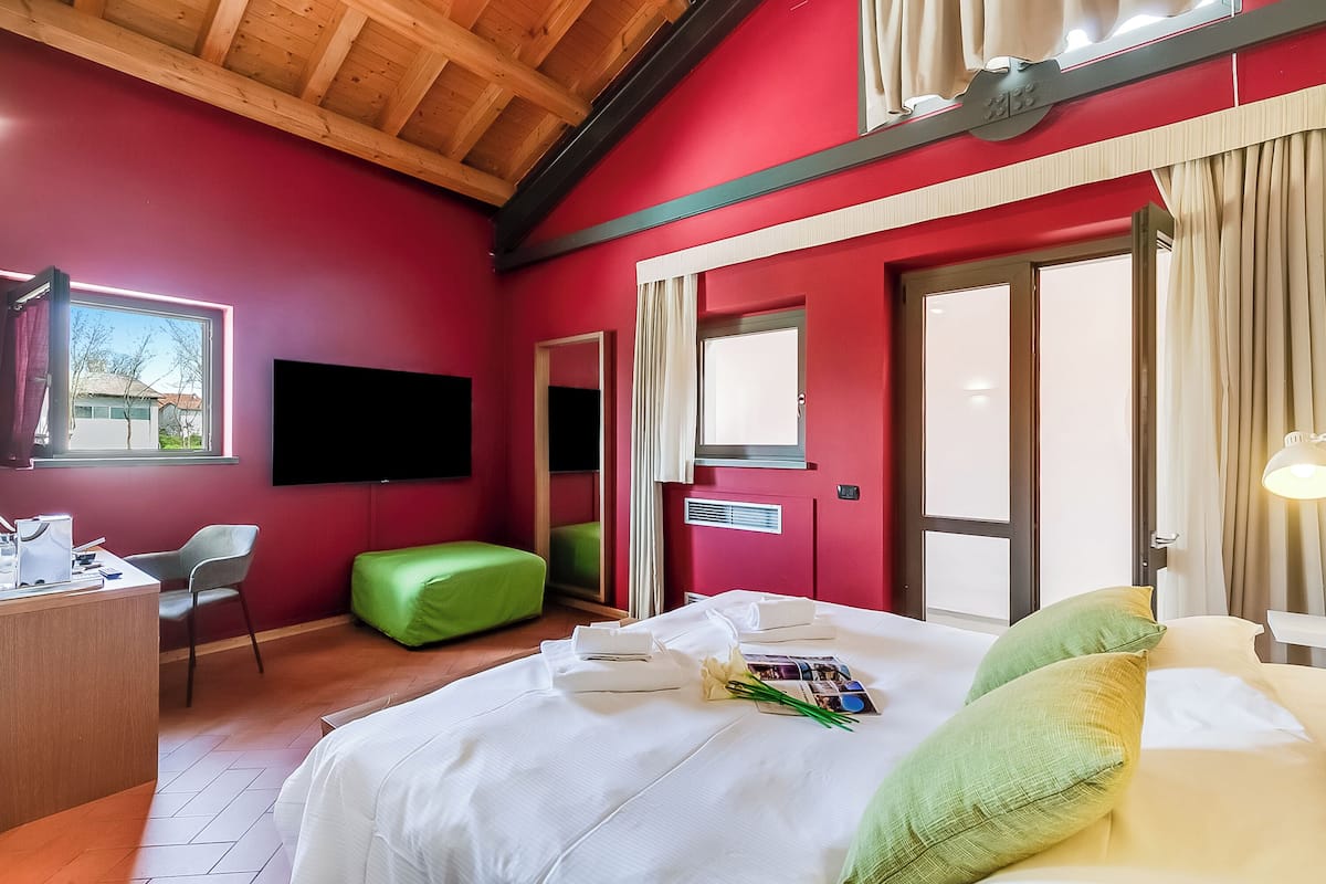 Habitación de hotel con pared roja, techo de madera, cama con ropa blanca, reposapiés verde, escritorio con silla y televisor de pantalla plana montado en la pared.